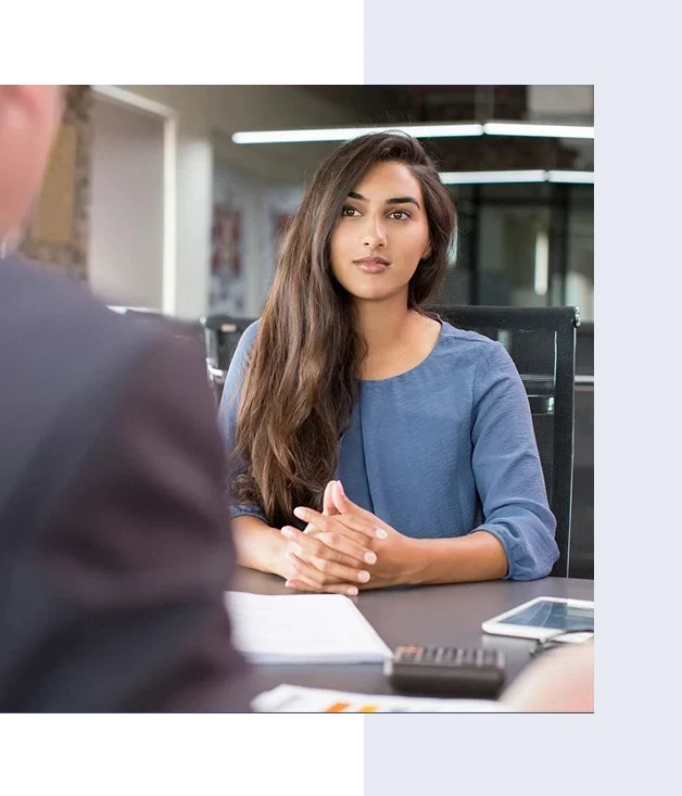 A woman with long hair sits at a table, listening attentively during a meeting. She wears a blue top and has a serious expression. A man is partially visible in the foreground.