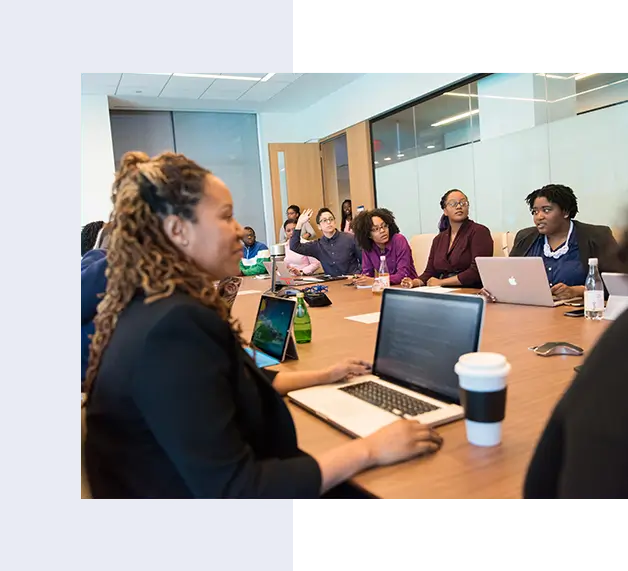 A group of people seated around a conference table with laptops and notebooks in a meeting room. Some are taking notes while one person is speaking.