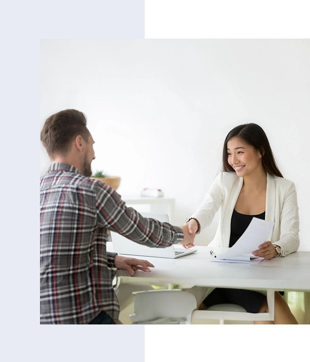 A man and woman shaking hands across a desk in an office setting, the woman holding papers and smiling.