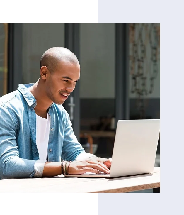 A person is sitting outdoors at a table, smiling and typing on a laptop. They are wearing a denim shirt over a white t-shirt.