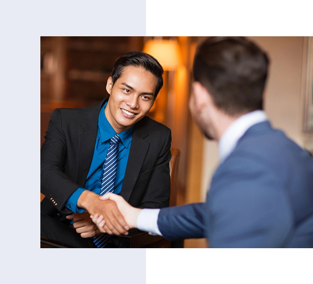 Two men in business attire are shaking hands in a formal setting, one man facing the camera and smiling while the other has his back to the camera.