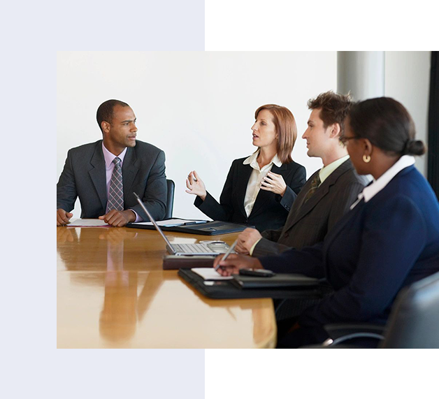 Four people in professional attire are seated around a conference table engaged in a discussion. One woman is talking while the others listen attentively. Laptops and documents are on the table.