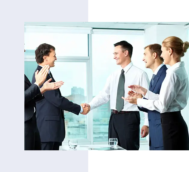 Four business professionals clapping while two men in suits shake hands in an office setting.
